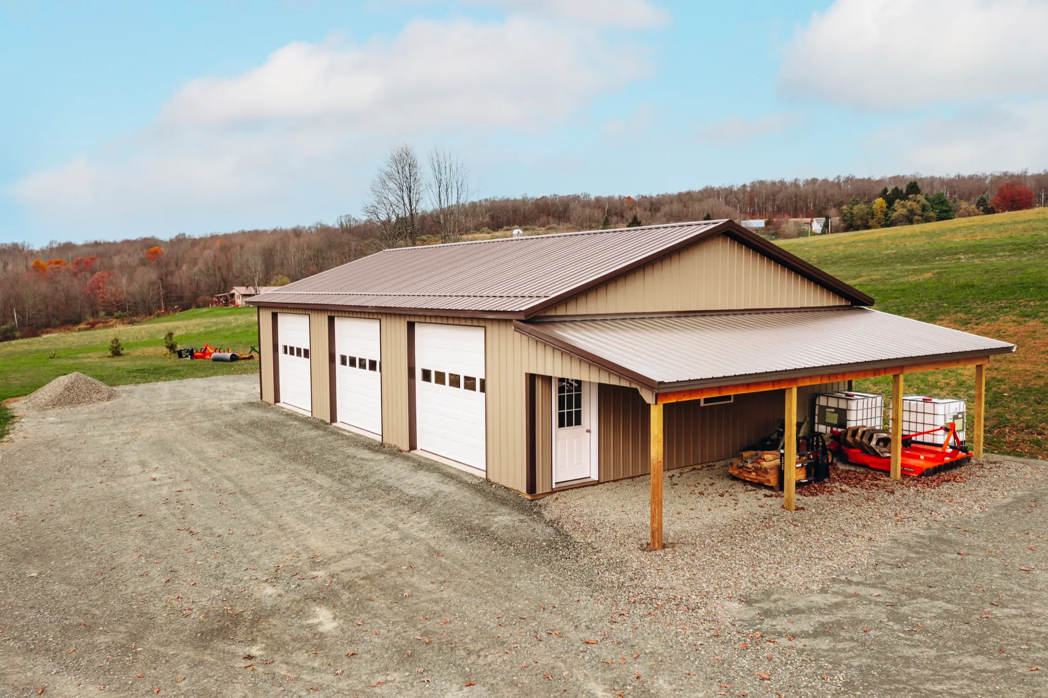 Aerial view of a tan post-frame building with a brown metal roof, three white overhead doors, and an open lean-to bay along the side sheltering equipment