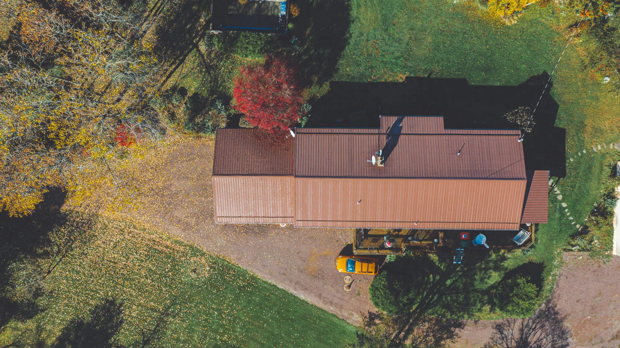 Overhead drone view looking straight down at a long building's new brown metal roof, ringed by autumn trees and a gravel drive