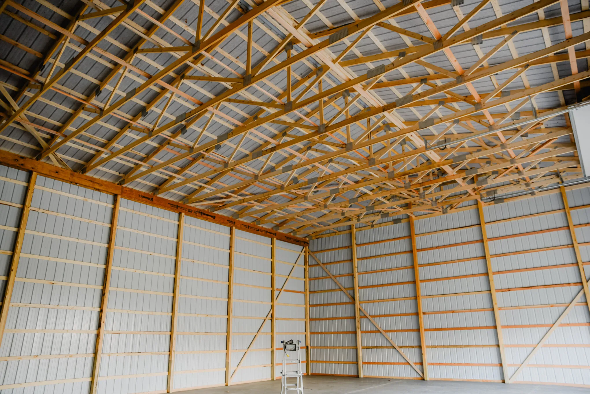 Inside an empty post-frame building looking up at long-span wooden roof trusses over white metal wall panels and a concrete floor