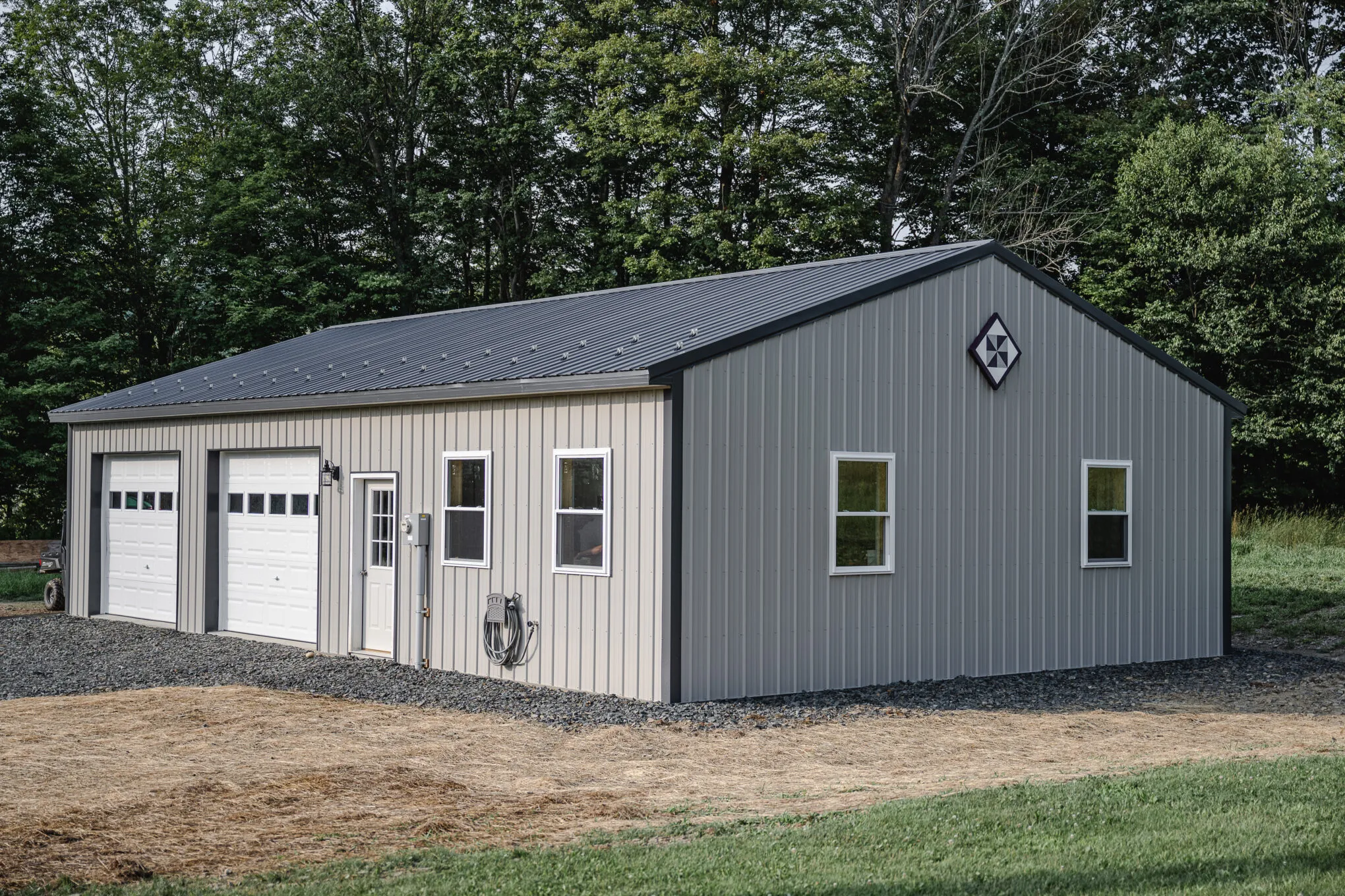 Post-frame garage with tan siding and a gray gable end, a charcoal metal roof, two white overhead doors, and a barn quilt on the gable