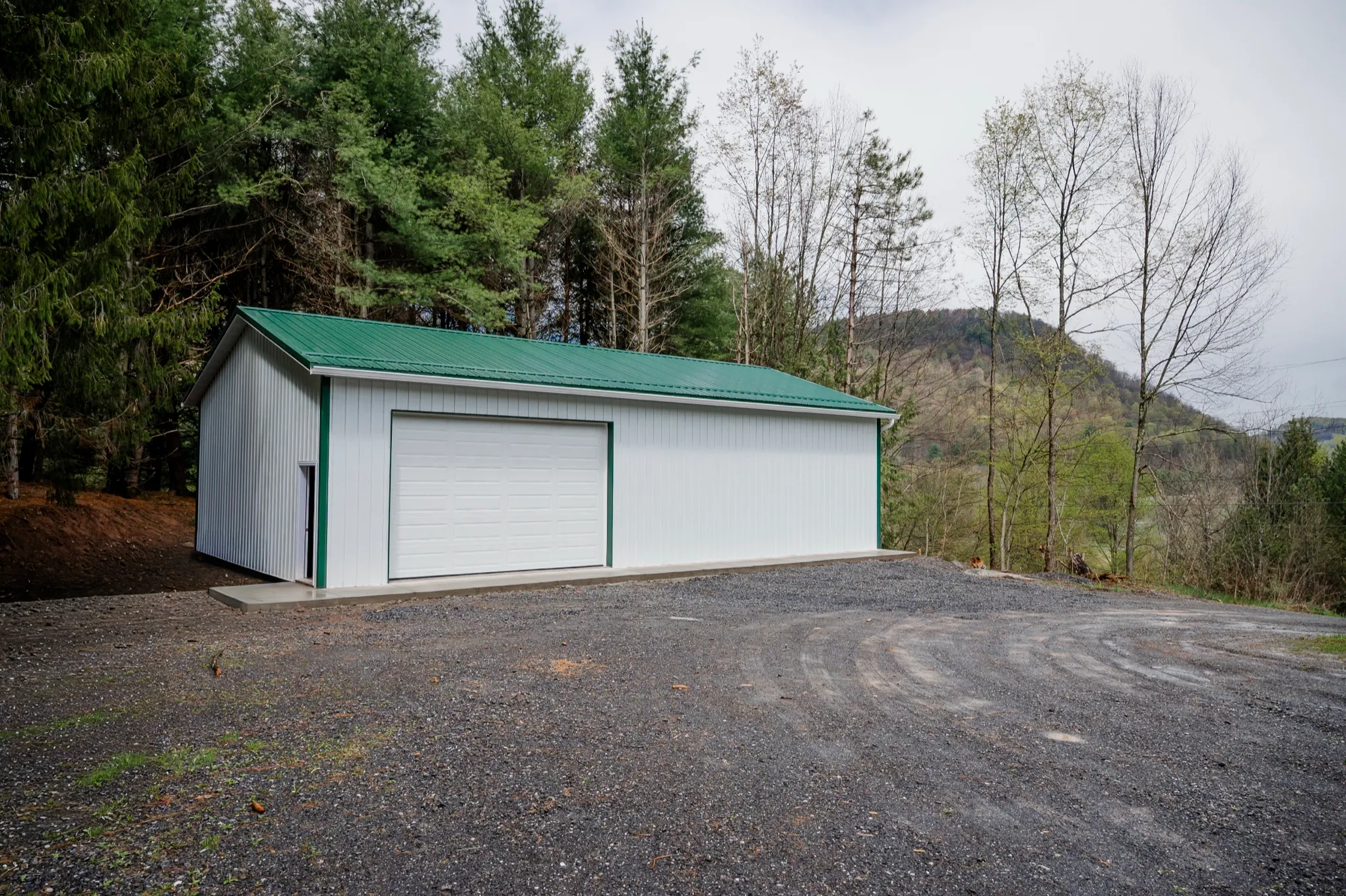 White post-frame garage with a green metal roof and green corner trim, a white overhead door and a white entry door, on a gravel pad above a wooded valley