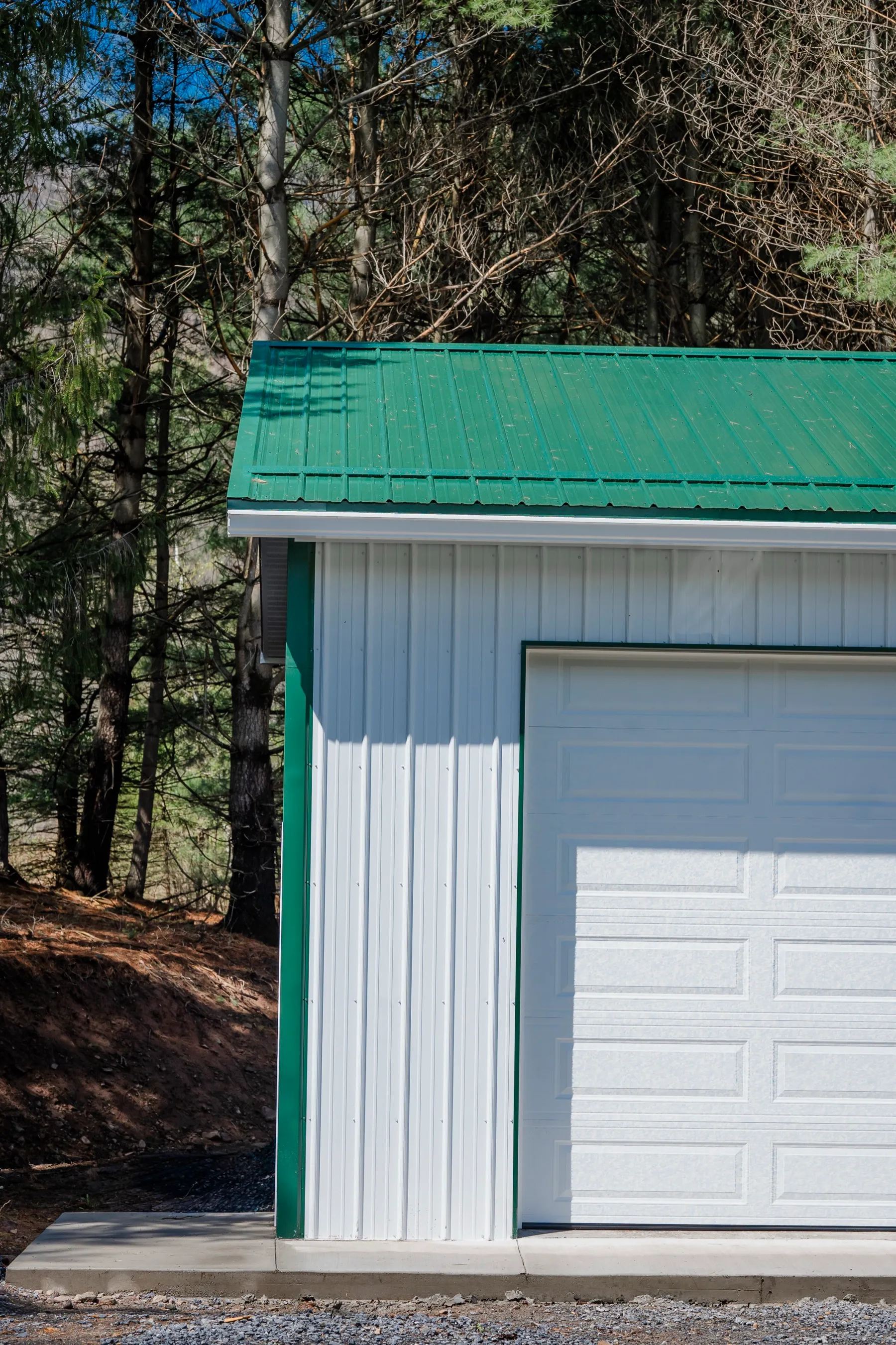 Close view of the corner of a white post-frame garage, showing the green metal roof, green trim, and a white overhead door against vertical white siding