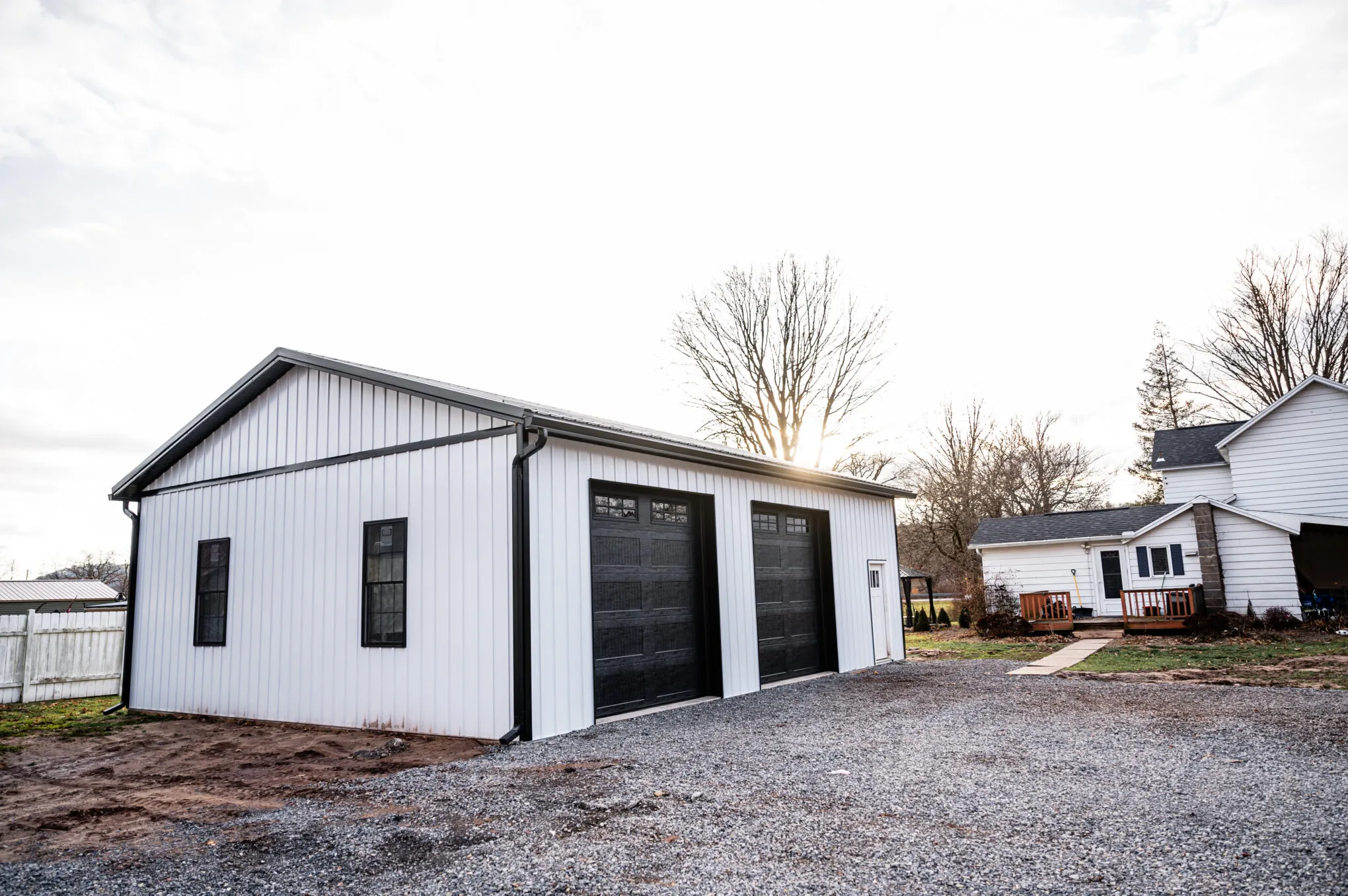 White post-frame garage with black trim, two black wood-grain overhead doors and black-framed windows on the gable end, beside a house