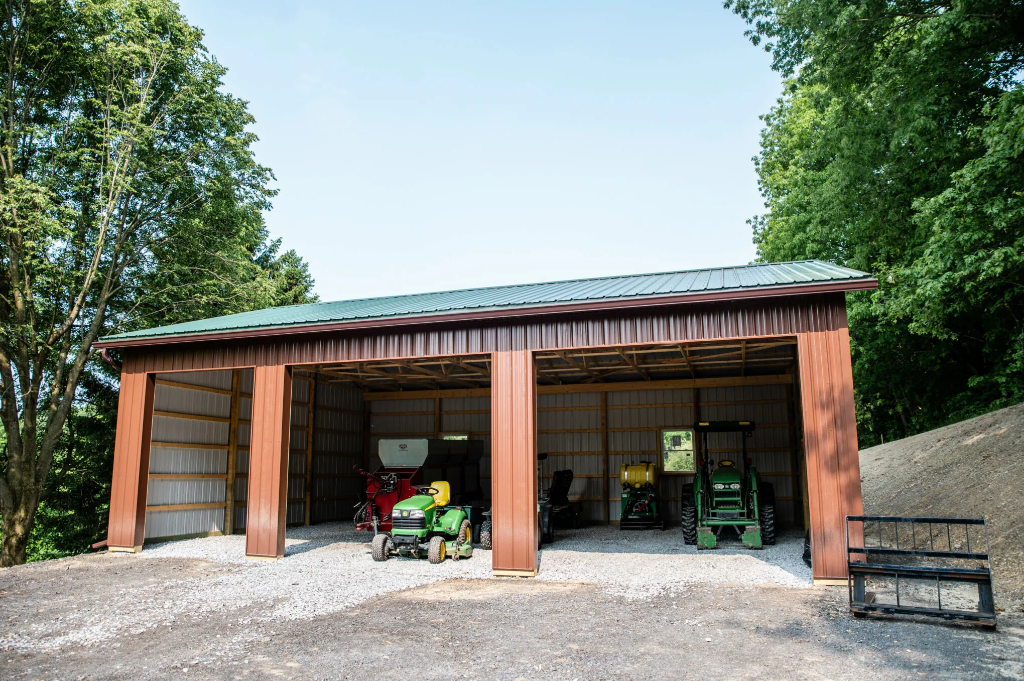 A rust-red post-frame equipment building with a green metal roof and open bays, with tractors parked inside