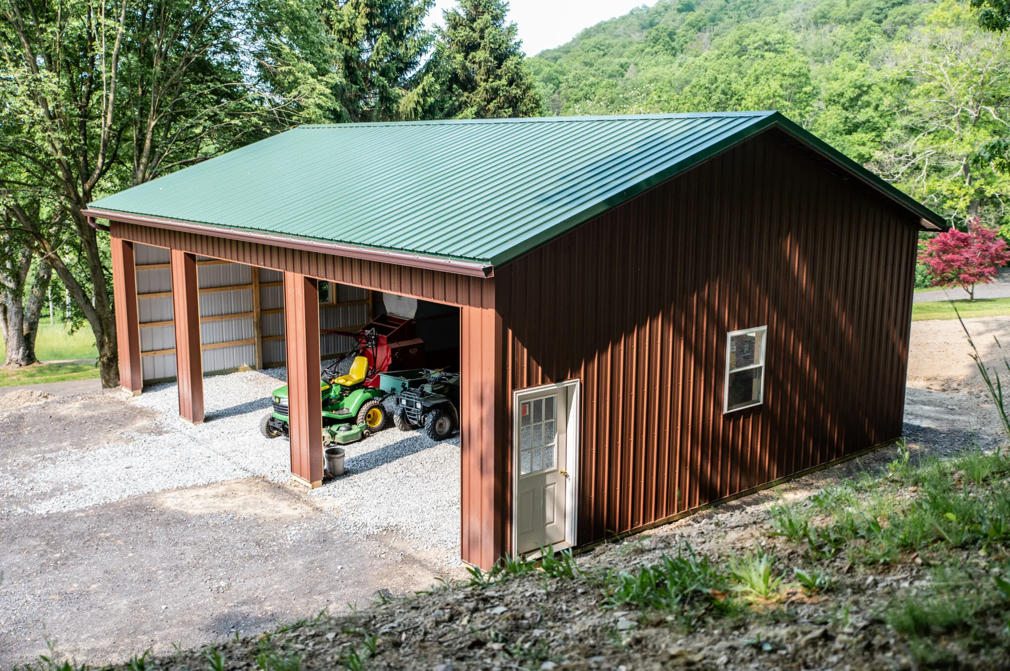 Brown post-frame building with a green metal roof and an open equipment bay sheltering a riding mower and an ATV