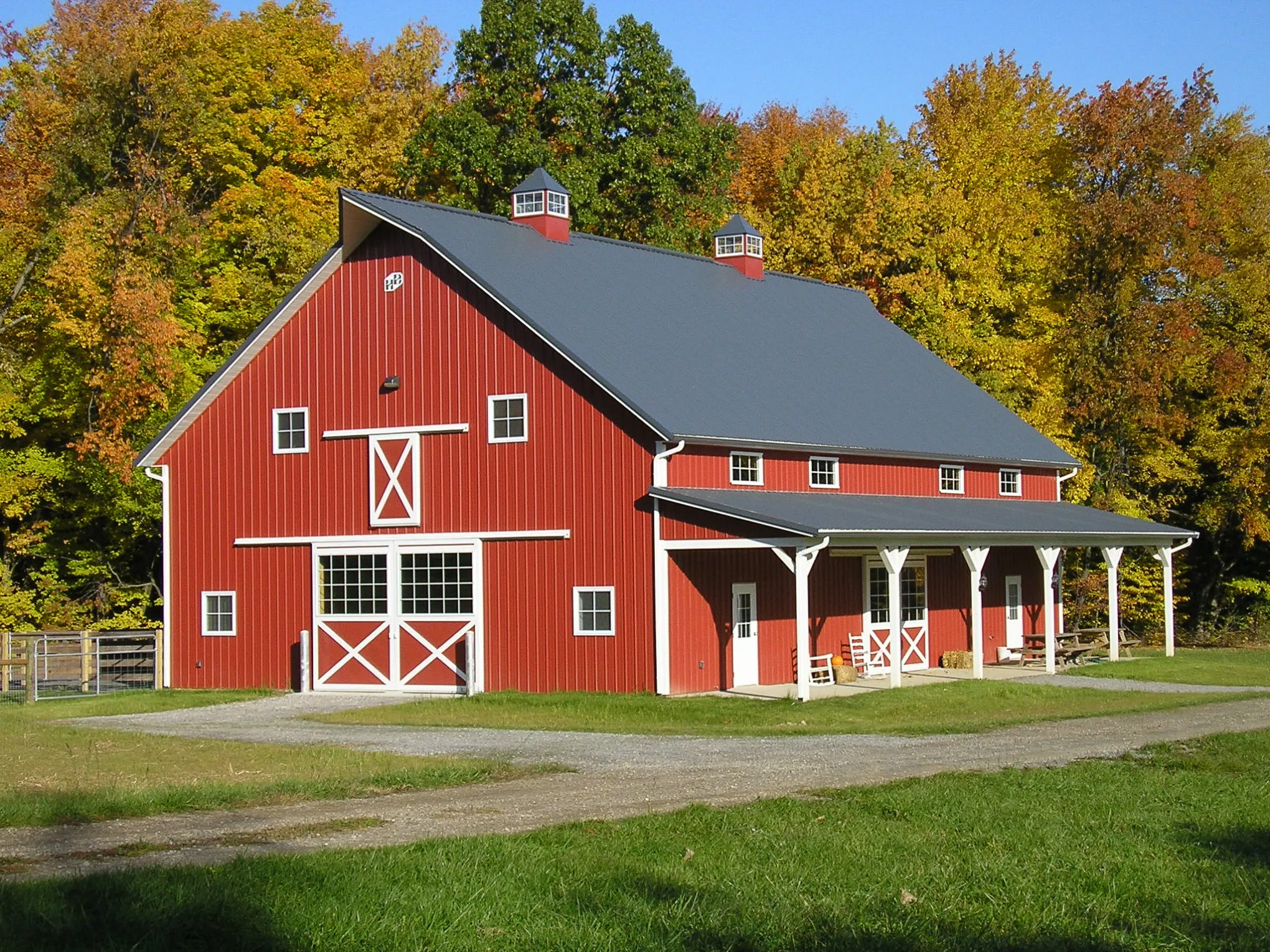 Large red barn with a charcoal metal roof, two cupolas, white cross-buck doors, and a covered porch along the side, framed by autumn trees