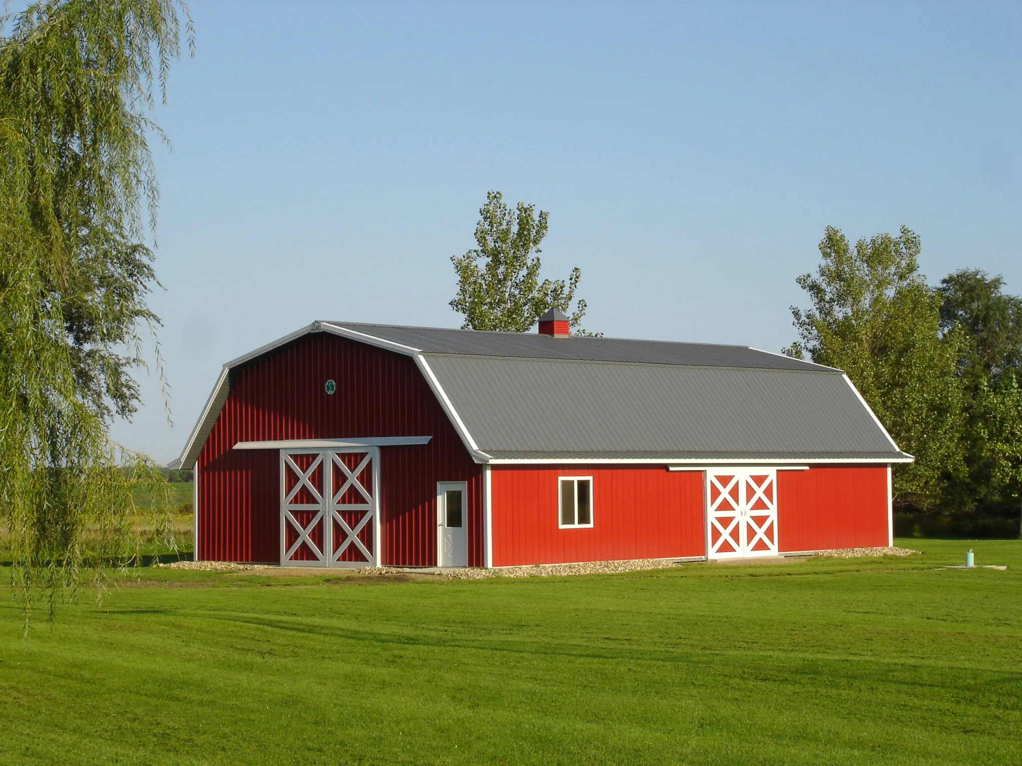 A red gambrel-roof barn with white cross-buck sliding doors and a gray metal roof, standing on a mown lawn