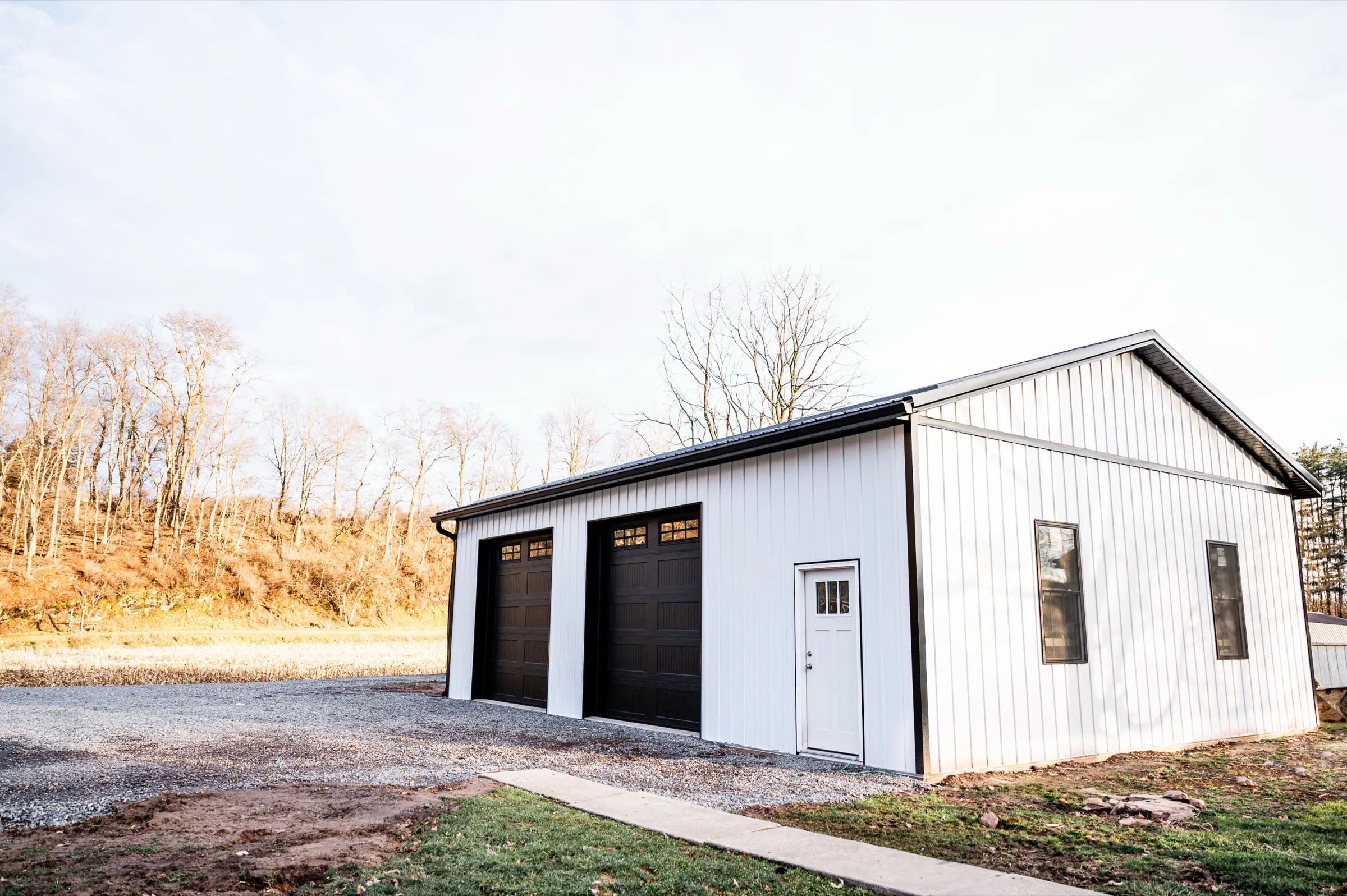 A white post-frame garage with two dark overhead doors and a white entry door, on a gravel drive with a wooded hillside behind
