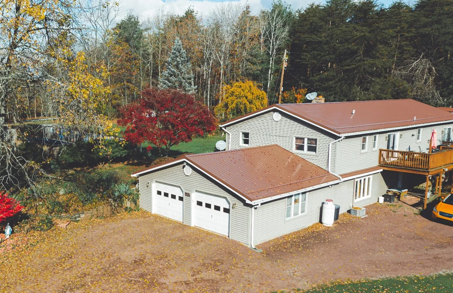 A two-storey cream-sided house and its attached two-car garage, both under a new brown metal roof, photographed from above among autumn trees